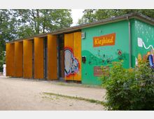 Grünes Gebäude mit gelber Holzverkleidung an einer Seite und Wandmalereien sowie der Aufschrift „Kiezkind Berlin“ an der anderen Seite; im Vordergrund ein sandiger Weg, umgeben von Bäumen und Sträuchern.