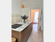 Moderne Küche mit einer Kücheninsel aus Holz und einer Arbeitsplatte aus weißem Marmor, darauf steht eine Vase mit lilafarbenen Blumen. Im Hintergrund sind weiße Wandschränke, ein Fliesenwandbereich und eine geöffnete Tür, die zu einem beleuchteten Flur führt.