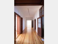 Ein langer, schmaler Flur mit hellen Holzdielen, weißen Wänden und hohen Türen aus dunklem Holz, die in angrenzende Räume führen. Am Ende des Flurs befindet sich eine offene Tür, durch die helles Tageslicht einfällt.