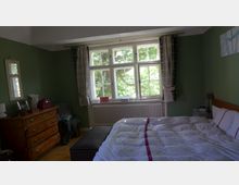 Schlafzimmer mit grünen Wänden, einem großen Fenster mit weißen Sprossenrahmen und karierten Vorhängen. Im Raum gibt es ein Doppelbett mit gemusterter Bettwäsche, eine Kommode aus Holz, einen kleinen Nachttisch sowie einen Heizkörper unter dem Fenster.