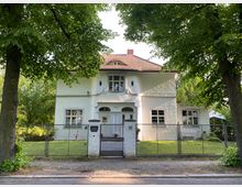 Zweistöckiges weißes Haus mit rotem Ziegeldach und verzierten Fenstern, umgeben von Bäumen und einer gepflegten Grünfläche. Vor dem Gebäude befindet sich ein Metallzaun mit einer Gartentür, die zur Straße führt.