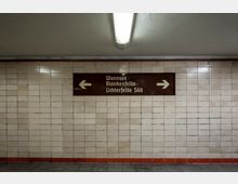 Unterführung mit beigefarbenen Fliesenwänden, einem braunen Hinweisschild mit weißer Schrift und Pfeilen, das Richtung "Wannsee", "Blankenfelde" und "Lichterfelde Süd" weist. Oben hängt eine rechteckige Deckenleuchte, der Boden ist mit grauen Fliesen bedeckt.