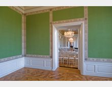 Ein prunkvoller Raum mit grünen Wänden, weißen Zierleisten und einem Parkettboden, durch eine geöffnete Tür ist ein großer Saal mit Reihen hellerer Stühle und glitzernden Kronleuchtern zu sehen. Im Hintergrund sind dekorative Stuckelemente und hohe Decken erkennbar.