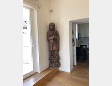 Holzstatue einer betenden Figur, die in einer hellen Ecke eines Raumes steht. Neben der Figur befindet sich eine Glastür mit Blick ins Freie, und im Hintergrund ist ein Durchgang zu einer modernen Küche erkennbar.