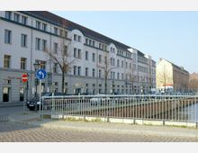 Straßenszene mit einem mehrstöckigen Gebäude im Hintergrund, das weiße Fassade und zahlreiche rechteckige Fenster hat. Im Vordergrund sind eine gepflasterte Straße, mehrere Verkehrszeichen, eine kleine Grünfläche mit einem Metallgeländer und ein flacher Wasserkanal sichtbar.