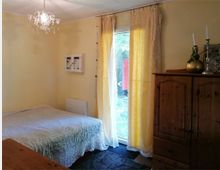 Kleines Schlafzimmer mit einem Bett auf der linken Seite, das mit einer hellen Überdecke bedeckt ist, und einem Kleiderschrank aus Holz auf der rechten Seite. Im Hintergrund befindet sich eine Glastür mit gelben Vorhängen, die zu einem Garten führt, und eine kleine Deckenleuchte hängt in der Mitte des Raums.