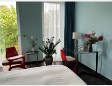 Modernes Schlafzimmer mit hellblauen Wänden, bodentiefen Fenstern mit transparenten Vorhängen und Blick ins Grüne. Im Raum stehen ein Glas-Schreibtisch mit Lampe und Blumen, rote Stühle, ein kleiner Beistelltisch und mehrere Zimmerpflanzen auf dunklem Holzboden.