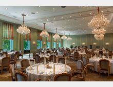 Eleganter Festsaal mit gedeckten, runden Tischen und gepolsterten Stühlen. Der Raum ist von großen Kronleuchtern beleuchtet, hat hohe Fenster mit gestreiften Vorhängen sowie hellgrüne Wände mit goldenen Zierleisten.