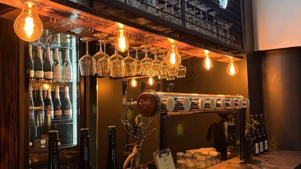 Innenansicht einer modernen Bar mit langer Theke aus Stein, hängenden Weingläsern und warmen Edison-Glühbirnen. Hinter der Theke stehen ein beleuchteter Getränkekühlschrank, Zapfhähne und ein Regal mit Neon-Schriftzug sowie gestapelten Gläsern.