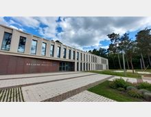 Modernes Gebäude des Rathauses Dallgow-Döberitz mit schlichter Fassade, großen Glasfenstern und flachem Dach, umgeben von einem gepflasterten Vorplatz und Grünflächen mit Bäumen im Hintergrund.