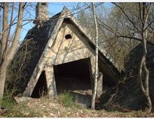 Verlassene Betonhütte mit dreieckigem Giebel und sichtbarer Beschädigung, umgeben von kahlen Bäumen und dichtem Unterholz in einem bewaldeten Gebiet.