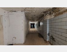 Langer, verlassener Flur mit einer stark beschädigten Decke und Wänden, teils verputzt und teils mit weißen Fliesen verkleidet. Der Boden ist mit einfachen Fliesen belegt, während Kabel und Rohre an der Decke freiliegen.