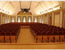 Großer Veranstaltungsraum mit einer holzgetäfelten Bühne und zahlreichen roten Stuhlreihen auf einem hellen Holzboden. Der Raum hat ein gewölbtes Deckengebälk, hohe Rundbogenfenster mit Vorhängen und helle Wände.