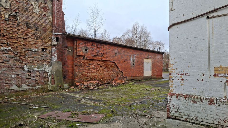 Verlassener Industriehof mit verwitterten Backsteinmauern, teils eingestürztem Mauerwerk und Schutt am Boden. Links und hinten sind rote Ziegelgebäude mit beschädigten Wänden zu sehen, rechts eine weiß gestrichene Ziegelwand mit abblätternder Farbe; der Boden ist rissig und von Moos überwachsen.