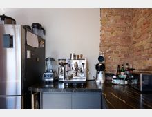 Kleine Küchenzeile mit einem Kühlschrank aus Edelstahl, einer schwarzen Arbeitsplatte und einer freiliegenden Ziegelwand. Auf der Arbeitsfläche befinden sich eine Espressomaschine, ein Mixer, eine Kaffeemühle, diverse Flaschen und Küchenutensilien.
