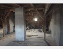 Dachboden eines älteren Gebäudes mit sichtbaren Holzbalken, rohen Wänden und einem kleinen Fenster, durch das Licht einfällt. Der Boden ist aus Holzdielen, teils beschädigt, und es liegen lose Bretter und Trümmer herum.