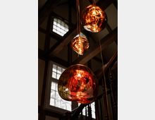 Drei hängende Glas-Pendelleuchten mit warmem, orangefarbenem Licht in einem Innenraum mit sichtbaren Holzbalken und großen, mehrteiligen Fenstern. Im Hintergrund ist ein Treppenaufgang mit dunklem Geländer zu sehen.