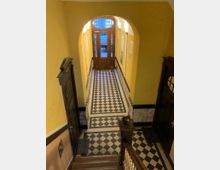 Treppenhaus mit gelber Wandgestaltung, schwarz-weiß gemustertem Fliesenboden und einer Holztreppe im Vordergrund, im Hintergrund eine verglaste Holztür mit ovalem Oberlicht.