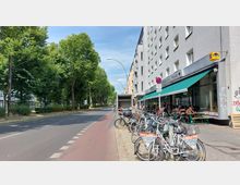 Eine städtische Straßenszene mit einem grau verputzten Gebäude auf der rechten Seite, das ein Café mit grüner Markise und Außenbestuhlung beherbergt. Links vom Gehweg verläuft eine breite Straße mit markiertem Fahrradweg, gesäumt von Bäumen und Büschen. Fahrräder stehen geparkt auf dem Gehweg, teils mit "Rent a Bike"-Schildern versehen.