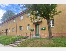 Zweistöckiges Wohngebäude mit gelb-beiger Fassade und roten Ziegelsteinen im unteren Bereich. Die Haustür ist grün mit zwei Briefkästen daneben, ein Baum und eine kleine Treppe befinden sich im Vordergrund.