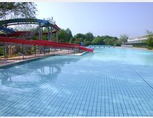 Ein Freibad mit einem großen, rechteckigen Becken und einer roten Rutsche, die sich geschwungen über mehrere Metallstützen erstreckt. Im Hintergrund sind grüne Bäume und ein Gebäude mit Glasfassade zu sehen.