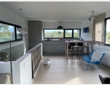 Helles, modernes Wohnzimmer mit offenen Küchenbereich. Der Raum verfügt über große Fenster mit Blick ins Grüne, eine graue Einbauküche, eine Holztheke mit Barhockern und eine Sitzgelegenheit mit blauem Stuhl.