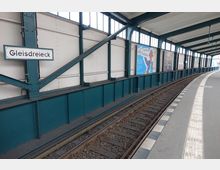 Überdachter U-Bahnhof mit Stahlkonstruktion, benannt "Gleisdreieck", mit gebogenem Bahnsteig, Gleisen und hellen Wänden; Werbeplakate sind entlang der Wand angebracht.