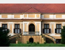 Fassade eines historischen Gebäudes mit gelb-beiger Putzfassade, barocken Verzierungen und mehreren symmetrischen Fenstern mit dunklen Holzläden. Eine zentrale Balustrade führt über gebogene Treppen zum Eingangsbereich mit einem großen Rundbogenfenster.