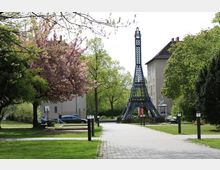 Ein kleiner Park mit gepflastertem Weg, umgeben von grünen Rasenflächen und blühenden Bäumen, darunter ein rosa blühender Baum. Im Hintergrund steht eine metallene Nachbildung des Eiffelturms, neben einem Wohnhaus.