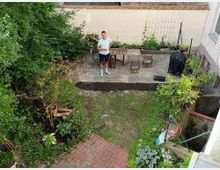 Ein kleiner Hinterhof mit einer gepflasterten Terrasse, auf der Gartenmöbel stehen, sowie einem Grill, der mit einer Abdeckung geschützt ist. Der Hof ist von Beeten mit Pflanzen und Büschen umgeben, und in der Mitte befindet sich ein teils mit Gras bewachsener Bereich.