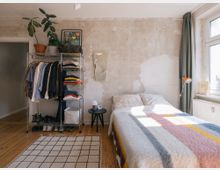 Schlafzimmer mit hellem Holzboden, einer abgenutzten, teilweise unverputzten Wand, einem niedrigen Bett mit bunter Decke, und einer kleinen schwarzen Nachttischlampe. Links steht ein offenes Kleiderregal mit Kleidung, Schuhen und Pflanzen, rechts ein großes Fenster mit grünen Vorhängen.