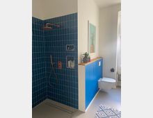 Modernes Badezimmer mit einer Dusche, die mit blauen Fliesen und einer Kupferarmatur gestaltet ist. Neben der Dusche befindet sich eine halbhohe Wand in Blau, hinter der eine weiße Wandtoilette montiert ist; oberhalb der Wand steht ein kleines Pflanzenarrangement neben einem Bild mit einer Ananas.