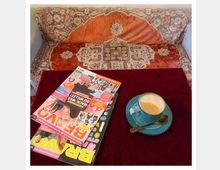 Ein kleines Tischarrangement mit einer roten Tischdecke, darauf eine Tasse Cappuccino mit Untertasse und Löffel in Blaugrün sowie eine bunte Zeitschrift. Im Hintergrund ein Sofa mit orientalisch gemustertem Überwurf in warmen Rot- und Beigetönen.