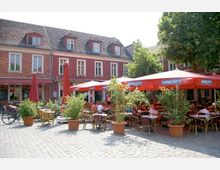 Marktplatz mit Kopfsteinpflaster, umgeben von roten Backsteingebäuden mit Satteldächern und Dachgauben. Im Vordergrund stehen Tische und Stühle eines Straßencafés unter roten Sonnenschirmen, dekoriert mit großen Pflanzen in Töpfen.