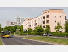 Mehrstöckige Wohngebäude mit hellen Fassaden stehen entlang einer Straße, umgeben von Grünflächen und Bäumen. Im Vordergrund fährt ein gelber Linienbus, daneben sind Verkehrszeichen und geparkte Autos zu sehen.