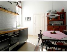 Helle Küche im klassischen Stil mit moderner Ausstattung, einschließlich schwarzer Unterschränke, einer Holz-Arbeitsplatte und weißer Fliesen im U-Bahn-Design an der Wand. Ein Esstisch mit rot-weiß kariertem Tischtuch und ein rustikales Holzbuffet stehen im Raum, geöffnet durch ein Fenster mit Holzjalousie.