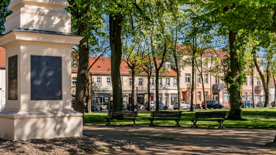 Tree-lined town square with a pale stone monument pedestal featuring dark plaques, benches along a paved path, and a grassy park area. Low historic buildings with red-tiled roofs and storefronts line the street in the background.