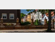 Reihen von Wohnhäusern in unterschiedlichen Farben und Baustilen, die hinter einer alten Backsteinmauer mit geschwungenen Formen und schmiedeeisernem Zaun stehen; im Vordergrund Gehweg und Baumstämme, im Hintergrund Bäume und blauer Himmel.