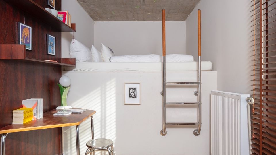 Compact loft bedroom with an elevated bed accessed by a metal ladder under an exposed concrete ceiling. The narrow room includes a built-in wood desk and shelving on one wall, beige carpet, and a large window with wooden blinds and a radiator beneath.