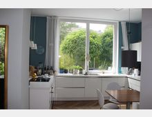 Moderne Einbauküche mit weißen Schränken, einer großen Fensterfront mit Blick auf grüne Bäume, und einem kleinen Esstisch mit zwei Stühlen. Im Vordergrund befindet sich ein Kochfeld, im Hintergrund sind Küchengeräte, Dekoration und Vorhänge sichtbar.