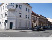 Straßenecke mit mehrstöckigen Wohngebäuden, darunter ein weißes Gebäude mit Balkongeländer und darüber ein Gebäude mit rot-braunen Fassadenelementen. Im Vordergrund eine Ampel, Verkehrsschilder sowie parkende und fahrende Autos auf der asphaltierten Straße.