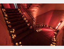 Treppenhaus mit metallenen Geländern, dekoriert mit zahlreichen leuchtenden LED-Kerzen auf den Treppenstufen und einem rötlich beleuchteten Ambiente. Die Wände und der Boden zeigen eine schlichte Gestaltung, ergänzt durch indirekte Beleuchtung.