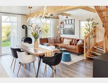 Helles Wohnzimmer mit moderner Einrichtung: Im Vordergrund ein Esstisch aus Holz mit schwarzen und weißen Stühlen, daneben eine braune Ledercouch mit Kissen, ein Kaminofen in der Ecke und eine Holztreppe im rechten Bildbereich. Große Fenster lassen viel Tageslicht in den Raum, im Hintergrund ist eine Grünfläche sichtbar.