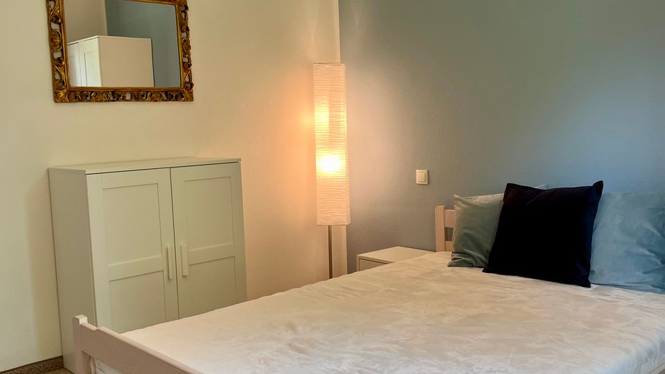 Schlafzimmer mit hellem Holzbett und weißer Bettwäsche, blaugrauer Akzentwand und Teppichboden. An der Wand hängen ein gerahmter Spiegel über einem weißen Schrank; neben dem Bett stehen ein kleiner Nachttisch und eine hohe Stehlampe, ergänzt durch eine schwarze Deckenleuchte.