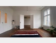 Ein heller Raum mit weißen Wänden und hohen Decken mit Stuckverzierungen. Zu sehen sind doppelflügelige Holztüren, ein großes Fenster mit weißen Vorhängen, ein Holzboden, ein roter Teppich, eine Schneiderpuppe und gestapelte Bücher sowie Pflanzen.