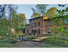 Backsteingebäude mit der Aufschrift „Annahütte“ in einer bewaldeten Umgebung, umgeben von jungen Bäumen und Pflanzen. Im Vordergrund sitzen zwei Personen auf Stühlen um eine Feuerstelle, und Holzstege führen über das Gelände.