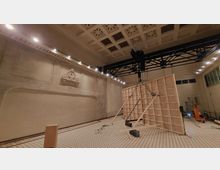 Großer Raum mit hoher Decke, verziert mit rechteckigen und durchbrochenen Mustern. Eine helle Wand mit einem kleinen Relief aus Stuck und eine Konstruktion aus Holzrahmen in der Raummitte; im Hintergrund sind moderne Beleuchtungselemente und technische Geräte zu sehen.