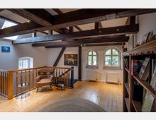 Ein gemütlicher Wohnbereich im Obergeschoss mit Holzbalkendecke, Holzboden und einer hölzernen Brüstung. Der Raum verfügt über drei abgerundete Fenster, einen Sessel mit kleinem Beistelltisch, Regale an der Seite und Dekorationen wie Gemälde und Bücher.