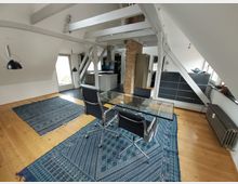 Ein helles Dachgeschosszimmer mit schrägen Wänden und sichtbaren weißen Holzbalken. Der Raum hat einen Holzboden, zwei blaue Teppiche, einen Glas-Schreibtisch mit schwarzen Stühlen sowie eine freistehende Backstein-Säule in der Mitte und eine halb offene Sicht auf eine angeschlossene Küche.