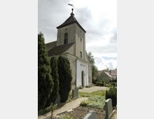 Kirche mit einem rechteckigen Turm und steilem Ziegeldach, umgeben von einem Friedhof mit Grabsteinen und gepflegten Grünanlagen. Im Hintergrund sind weitere Gebäude und ein bewölkter Himmel zu sehen.
